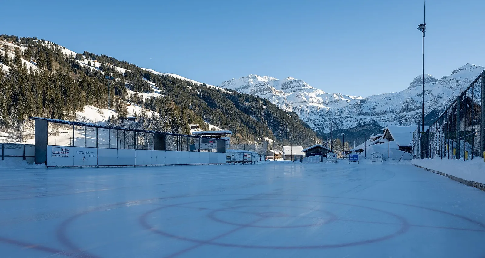 Die Kunsteisbahn Lenk im Simmental an einem sonnigen Wintertag, mit der bläulichen Eisfläche im Vordergrund und verschneiten Bergen und dem Tribünengebäude dahinter. – Kunsteisbahn Lenk (Sportanlage TEC)
