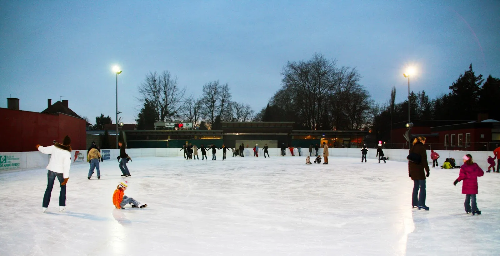 Skaters spread across the floodlit Leibnitz artificial-ice rink at dusk, with bare trees and lamp posts behind the boards. — Kunsteisbahn Marenzi