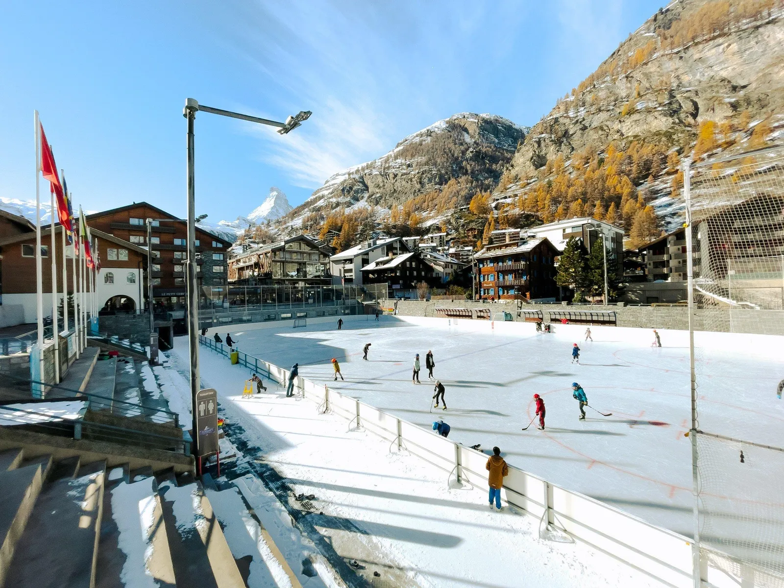 The Obere Matten ice rink in Zermatt on a sunny winter day, with skaters spread across the ice, a stand along the side and snowy mountains and hotels behind. — Kunst- und Natureisbahn Zermatt (Obere Matten)