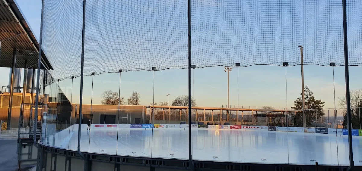 Das Freiluft-Eisfeld der Kunsteisbahn Oerlikon in Zürich in der Dämmerung, durch das Schutznetz gesehen, links die überdachte Halle – Kunsteisbahn Oerlikon