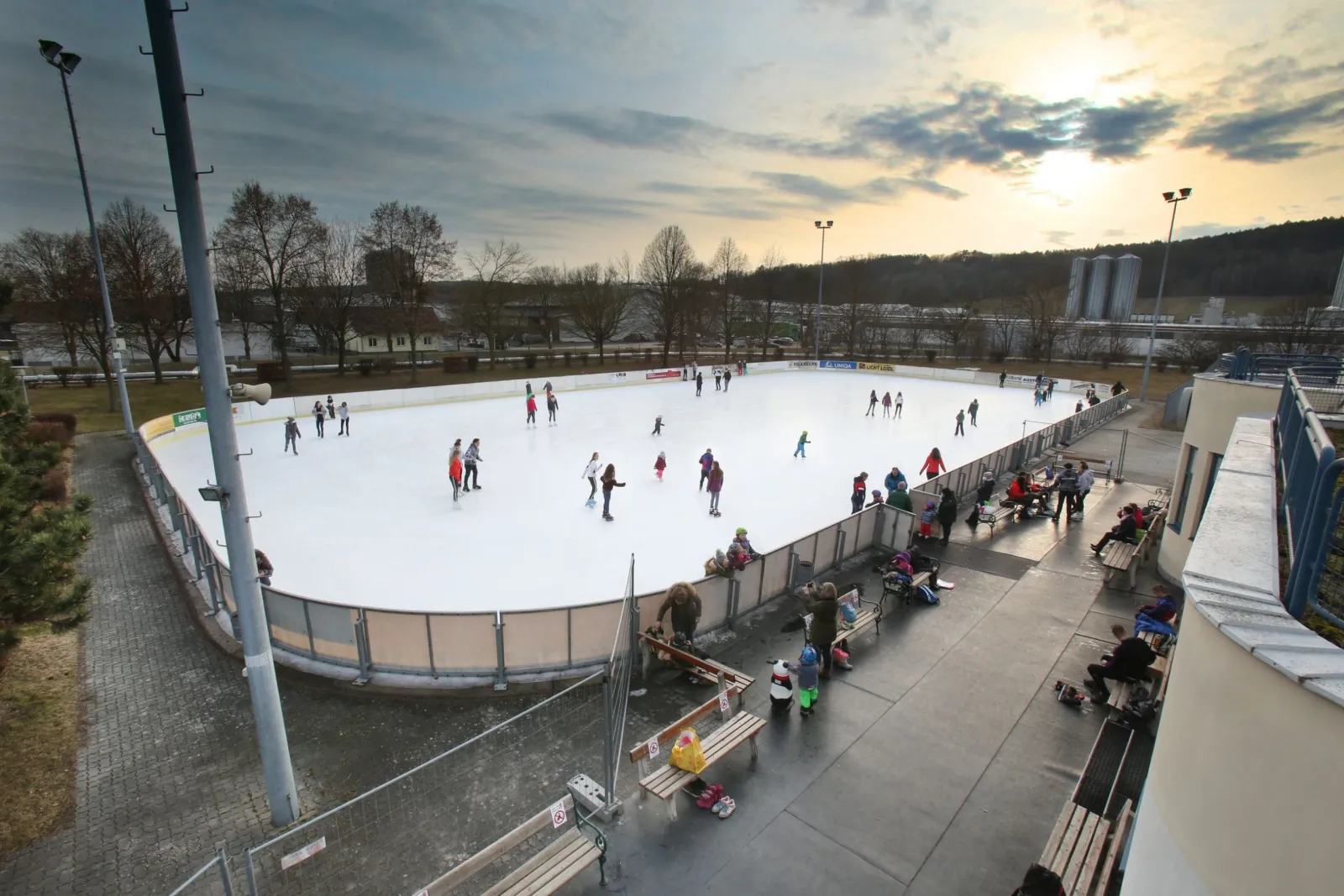 The open-air artificial ice rink in Pinkafeld at dusk, seen from above with skaters small across the ice and the boards around it — Kunsteisbahn Pinkafeld