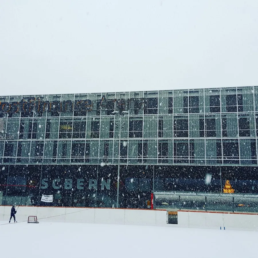 Das Freiluft-Eisfeld der PostFinance-Arena in Bern bei Schneefall, mit einem Tor auf dem Eis und der Arena dahinter – Kunsteisbahn PostFinance-Arena