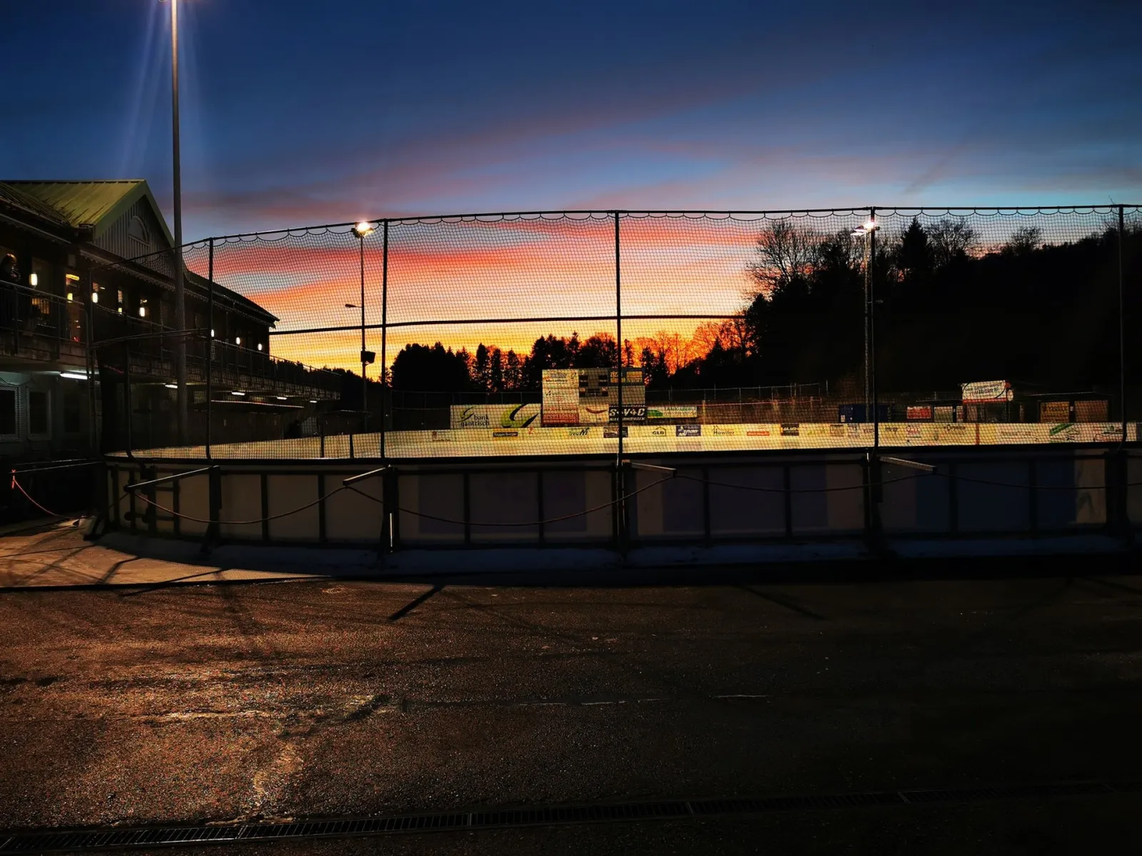 The outdoor rink at dusk behind its safety netting, with the clubhouse on the left, floodlights lit and an orange sky over the trees. — Kunsteisbahn Schwarzwasser