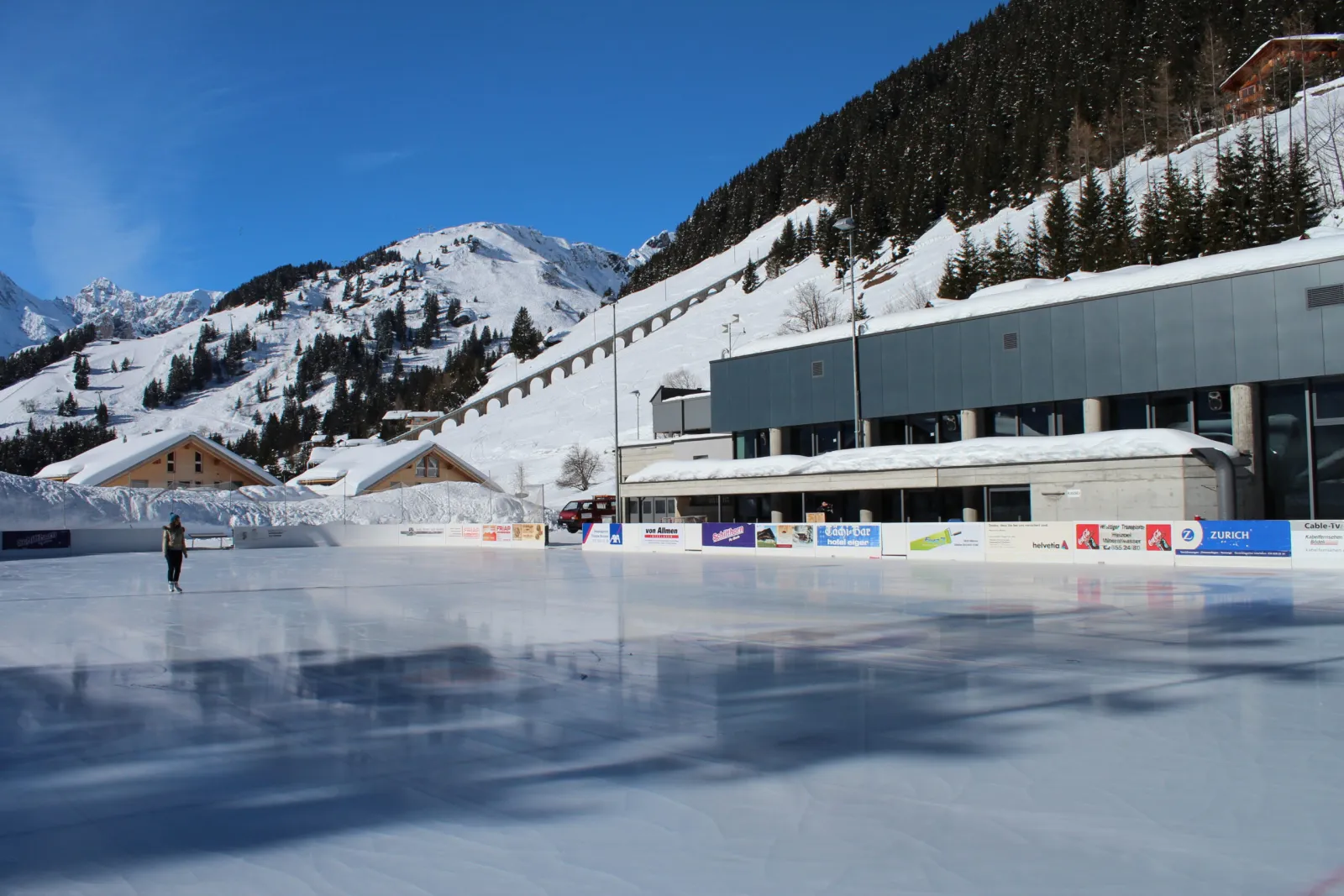 The open-air ice field in front of the Sportzentrum building, with the village and slopes behind — Kunsteisbahn Alpines Sportzentrum Mürren