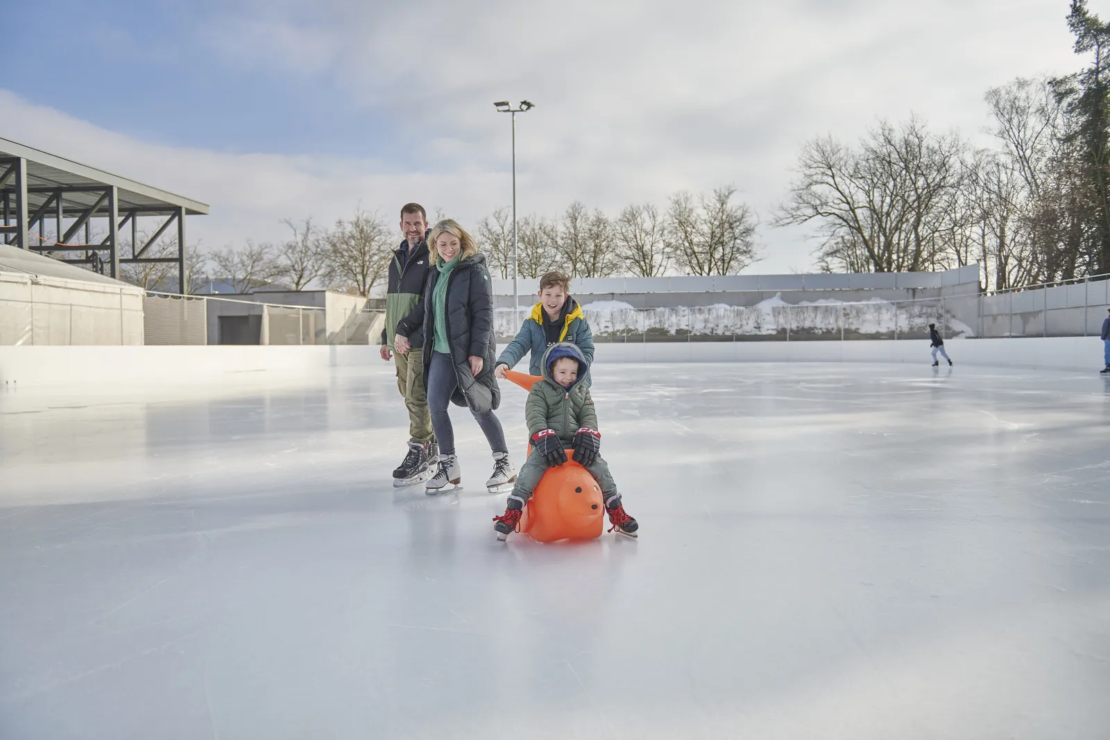 Familien laufen Schlittschuh auf der weiten Freiluft-Eisfläche im Tägi Wettingen; ein Elternteil schiebt ein Kind auf einem Sitz in Richtung Kamera, über ihnen die Stahldachkonstruktion. – Kunsteisbahn Tägerhard (Tägi)