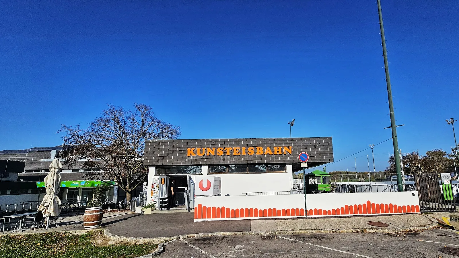 Das niedrige Gebäude der Kunsteisbahn Tulln mit orangefarbenen Namensbuchstaben am dunklen Dachrand, vom Parkplatz aus unter klarem blauem Himmel gesehen – Kunsteisbahn Tulln