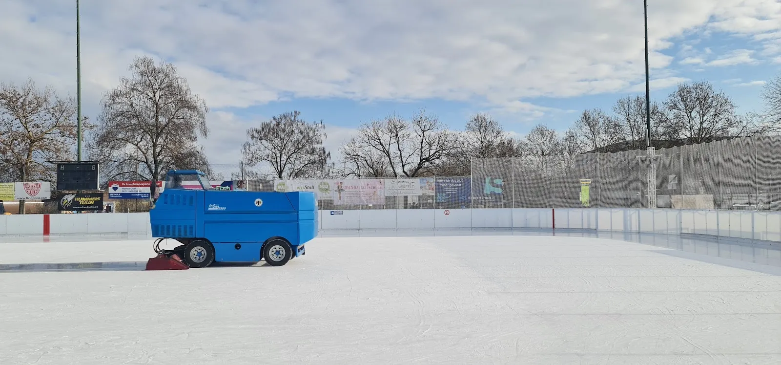 Das Freiluft-Eis der Kunsteisbahn Tulln mit der blauen Eismaschine bei der Arbeit auf der leeren Fläche und kahlen Bäumen hinter den Banden – Kunsteisbahn Tulln