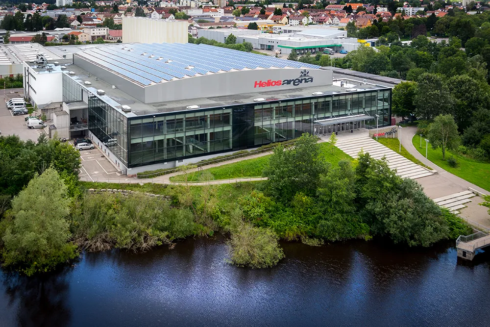 Aerial view of the Helios Arena in Villingen-Schwenningen, the glass-fronted arena with its long solar roof beside the river and park — Kunsteisbahn Villingen-Schwenningen