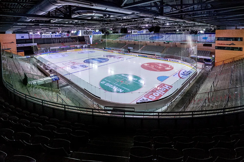 The empty ice of the arena in Villingen-Schwenningen seen from the upper tier, with sponsor logos on the ice and the dark seating bowl around it — Kunsteisbahn Villingen-Schwenningen