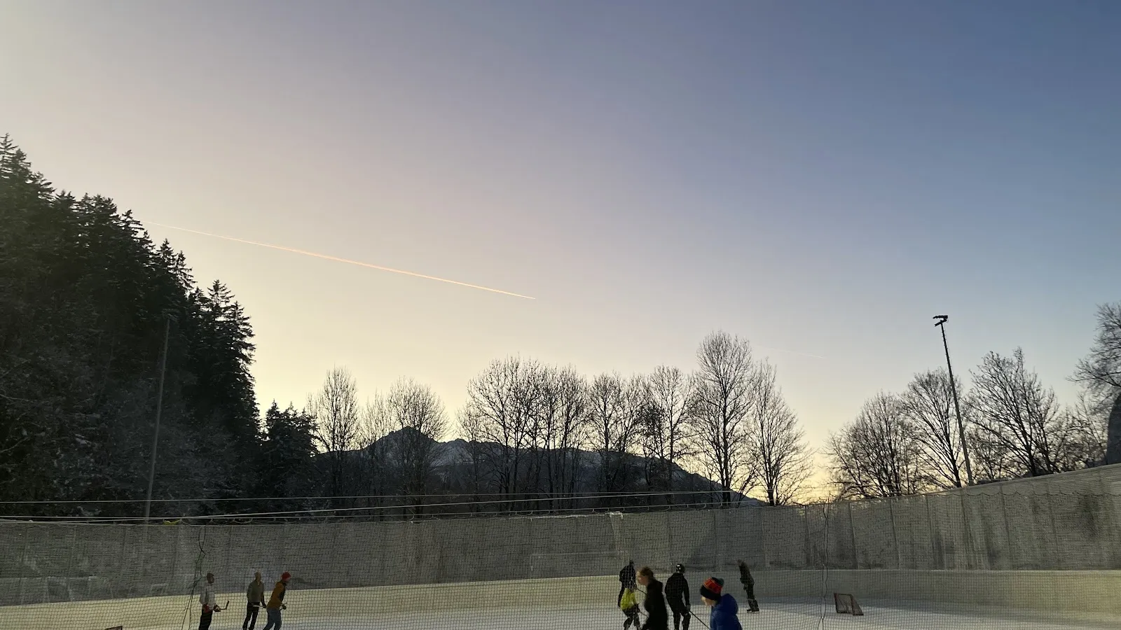 The open-air Eislaufplatz Völs at dusk with a few skaters on the ice and the wooded slope and floodlight masts behind the rink. — Kunsteisbahn Völs