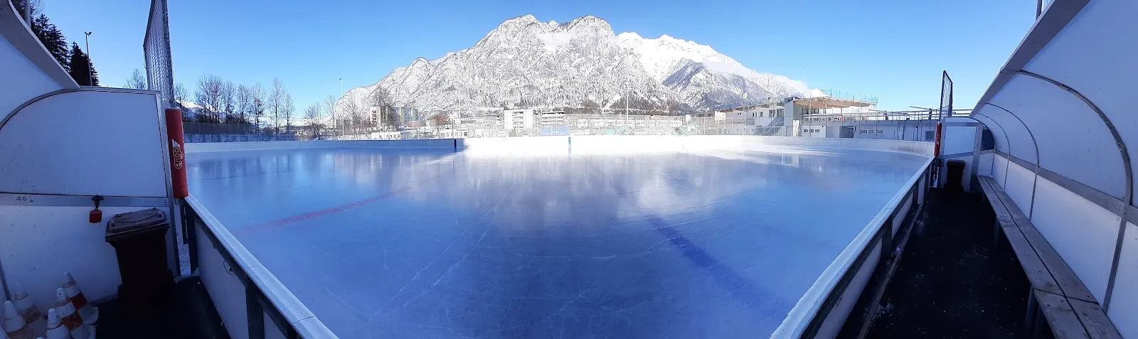 The outdoor ice sheet of the Eislaufplatz Völs, empty and frosted, seen from between the boards with snowy mountains behind (Maps listing: Eislaufplatz Völs) — Kunsteisbahn Völs