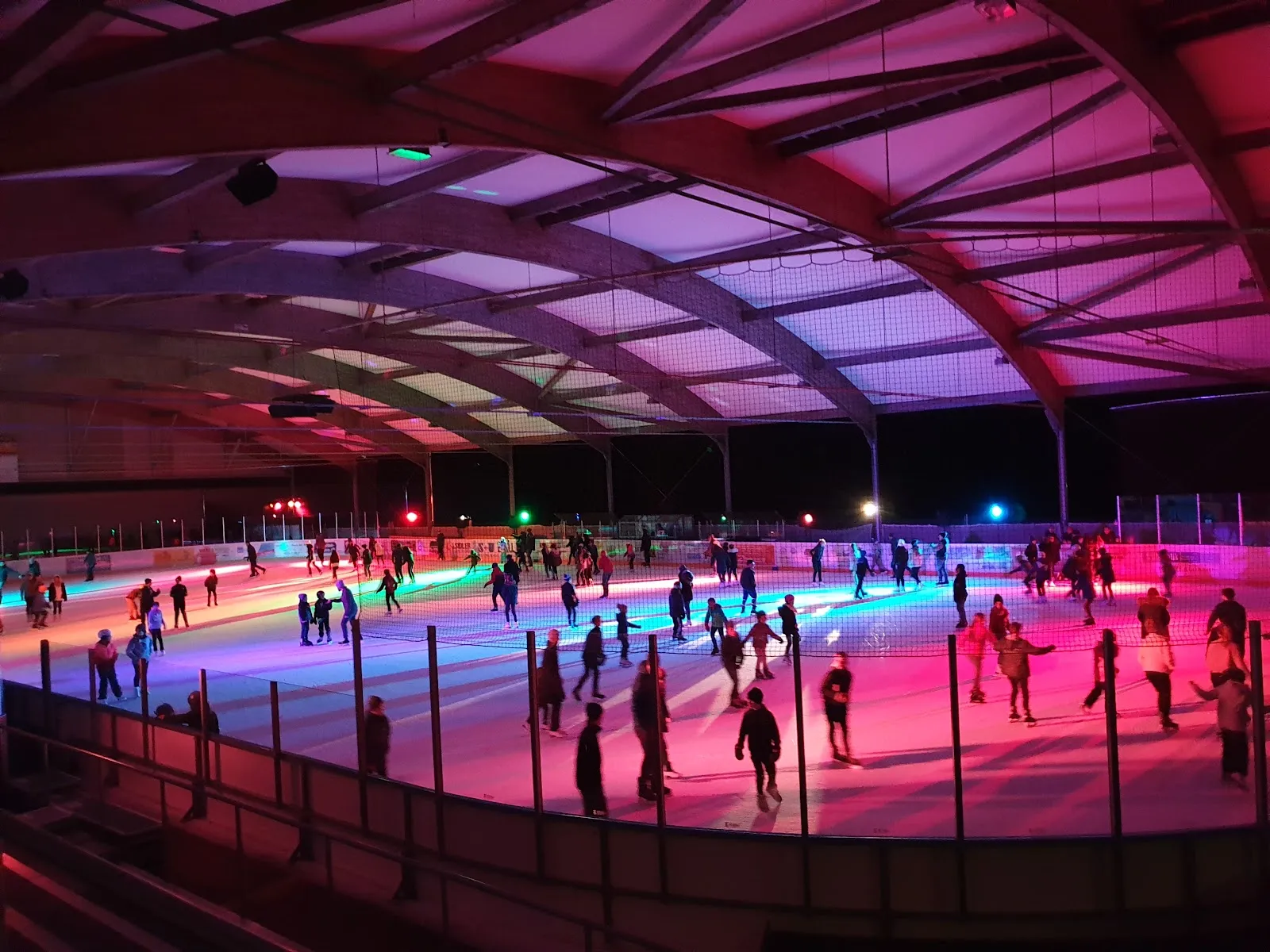 The covered rink lit pink and blue, the whole ice surface visible with the public skaters small in the distance and the arched roof above. — Kunsteisbahn Weihermatt