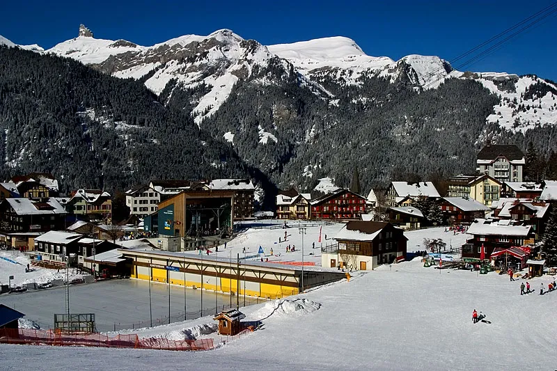 Die offene Eisbahn und ihr Clubhaus mit gelbem Dach am Rand von Wengen, dahinter die Jungfrau-Berge (Foto von 2004) – Kunsteisbahn Wengen