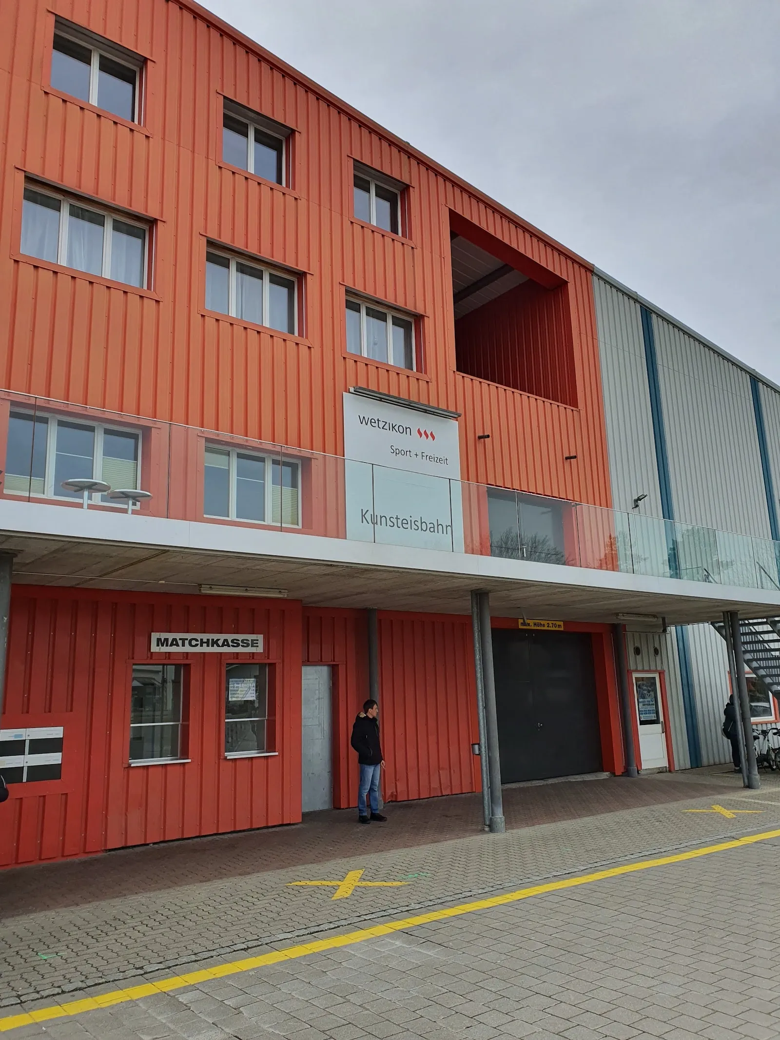 The red-clad front of the Kunsteisbahn Wetzikon with the Kunsteisbahn lettering above the Matchkasse ticket window and the covered walkway along the building. — Kunsteisbahn Wetzikon