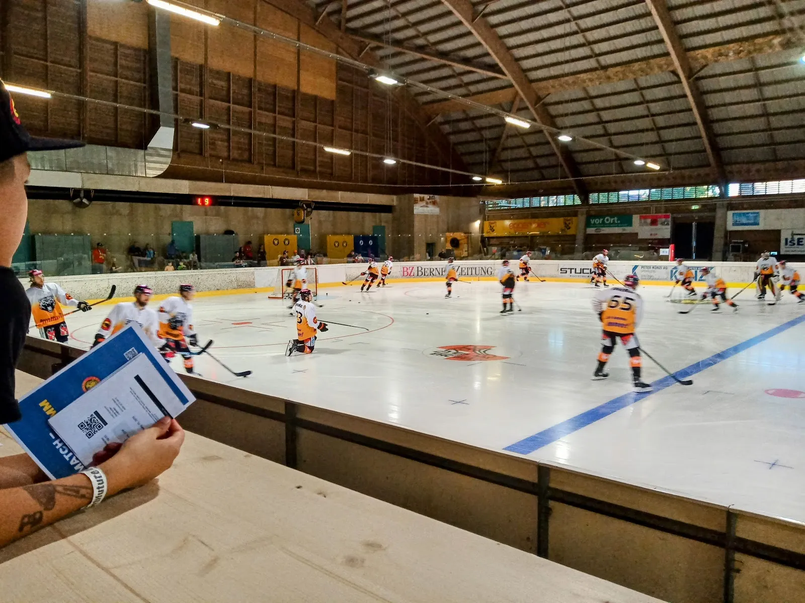 Die breite Eishalle mit einem Eishockeyspiel am anderen Ende, Banden und Tribünen; am Bildrand ist ein Arm zu sehen. – Kunsteisbahn Wislepark