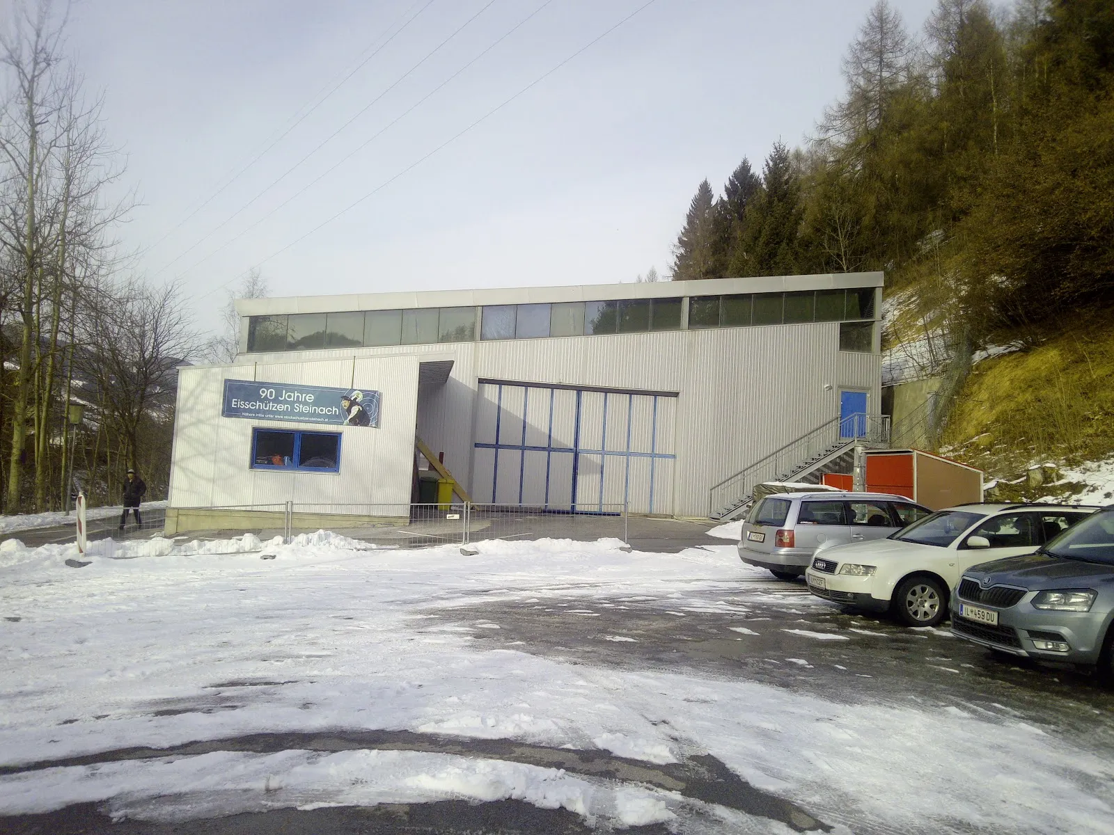 Die niedrige, grau verkleidete Kunsteis- und Mehrzweckhalle in Steinach am Brenner, über einen verschneiten Parkplatz gesehen, mit einem Banner an der Vorderwand – Kunsteis- und Mehrzweckhalle Steinach