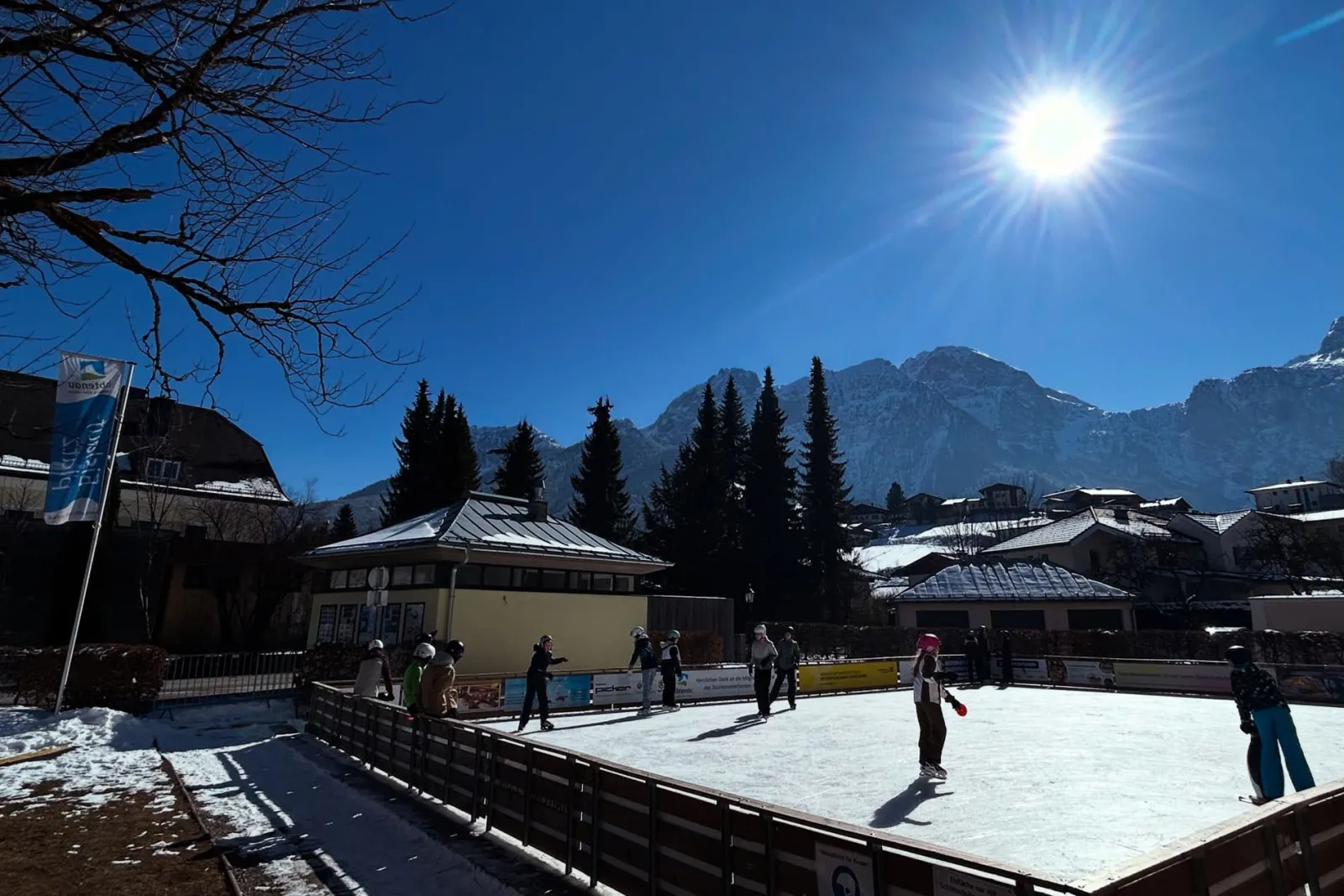 The boarded ice rink in the centre of Abtenau on a clear winter day, with a few skaters, low village buildings and mountains behind. — Kunsteislaufplatz Abtenau