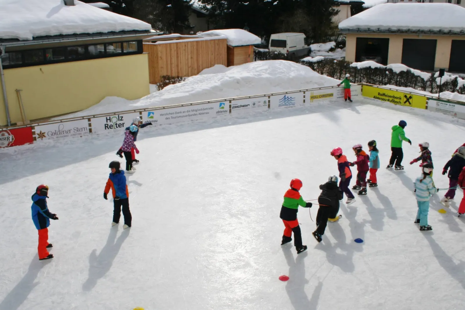 Children practising on the Abtenau ice rink between coloured cones, seen from above, with sponsor boards along the wooden rail and snowy roofs behind. — Kunsteislaufplatz Abtenau