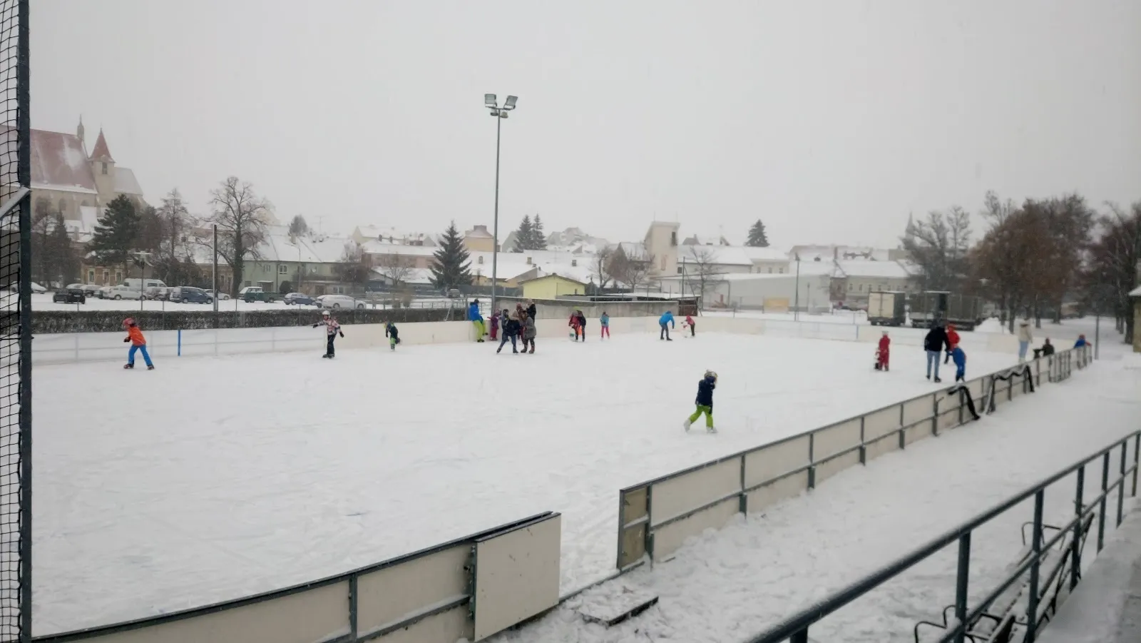 Der Freiluft-Kunsteislaufplatz in Eggenburg an einem verschneiten Tag, mit Läufern auf dem Eis und dem Zaun und niedrigen Gebäuden dahinter. – Kunsteislaufplatz Eggenburg