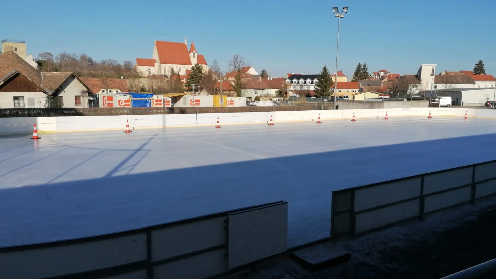 Die leere Freiluft-Kunsteisbahn in Eggenburg an einem sonnigen Tag, mit der Stadt und ihrer Kirche hinter den Banden – Kunsteislaufplatz Eggenburg