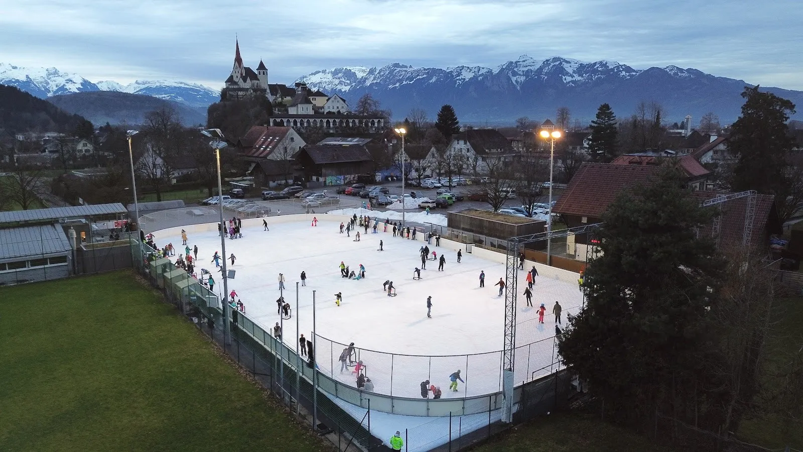 The open-air Gastra rink in Rankweil seen from above at dusk, skaters spread across the ice with the village, castle tower and mountains behind. — Kunsteislaufplatz Gastra Rankweil
