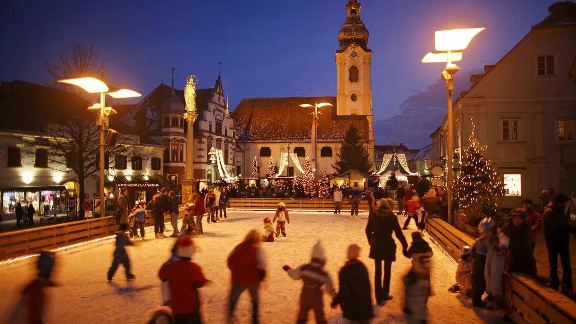 Skaters on the Christmas ice rink on Hartberg's town square at dusk, with the church tower, street lamps and festive lights behind. — Kunsteislaufplatz Hartberg