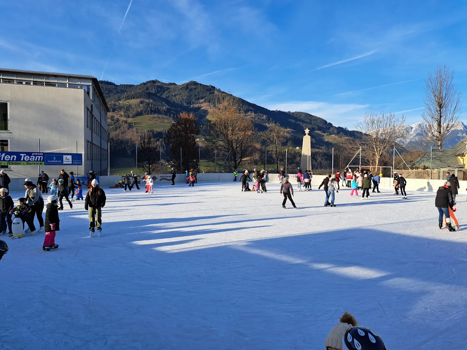 Ein sonniger Publikumslauf auf der Kunsteisbahn unter freiem Himmel in St. Johann im Pongau: Eisläufer verteilen sich auf der weiten Eisfläche, dahinter Banden, ein Schulgebäude und bewaldete Berge. – Kunsteislaufplatz St. Johann im Pongau