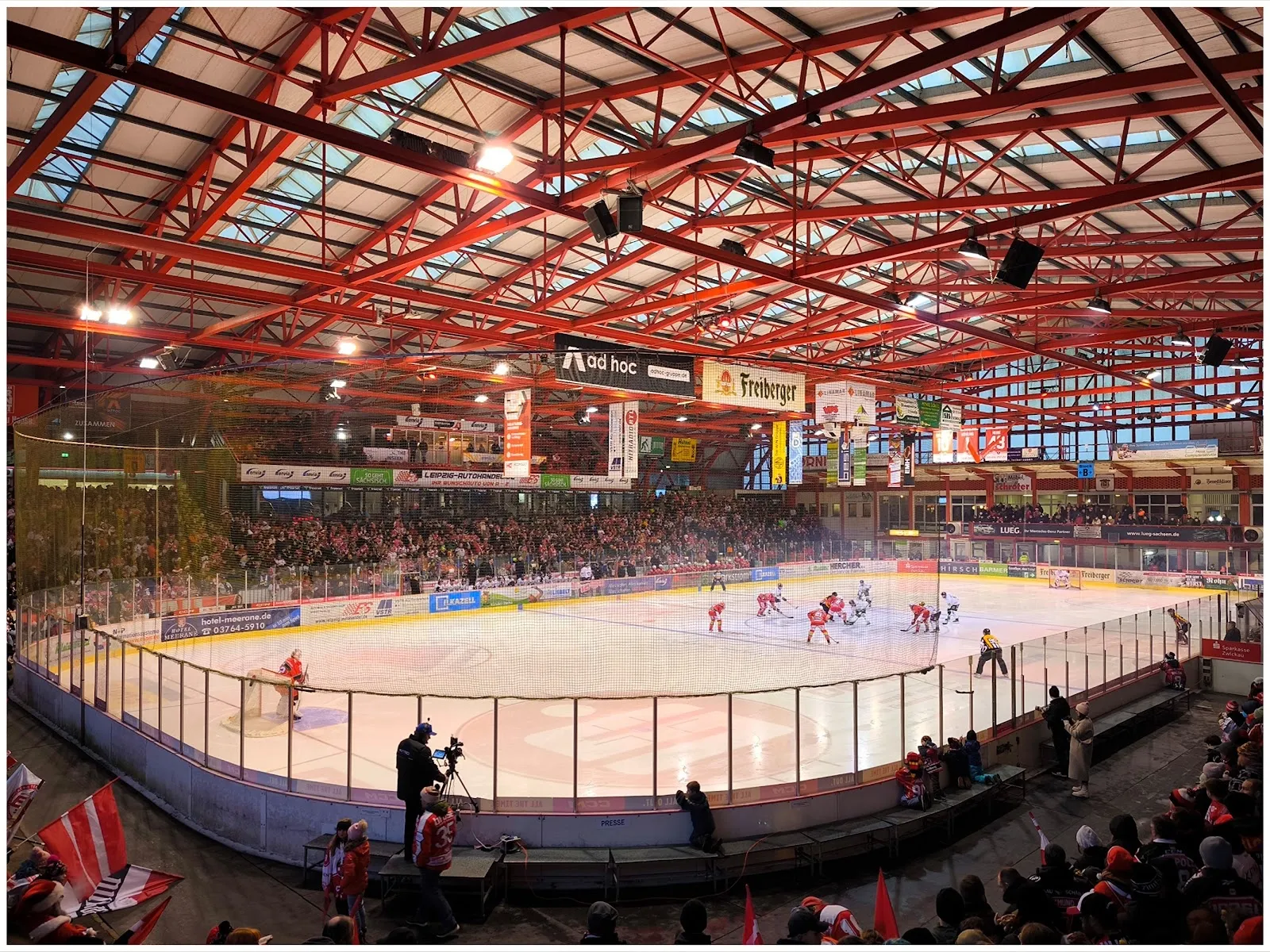 Das Eis des Kunsteisstadions im Sahnpark in Crimmitschau von den Rängen aus gesehen, unter dem roten Stahldach, mit Zuschauern auf den Rängen rund um die Fläche. – Kunsteisstadion im Sahnpark