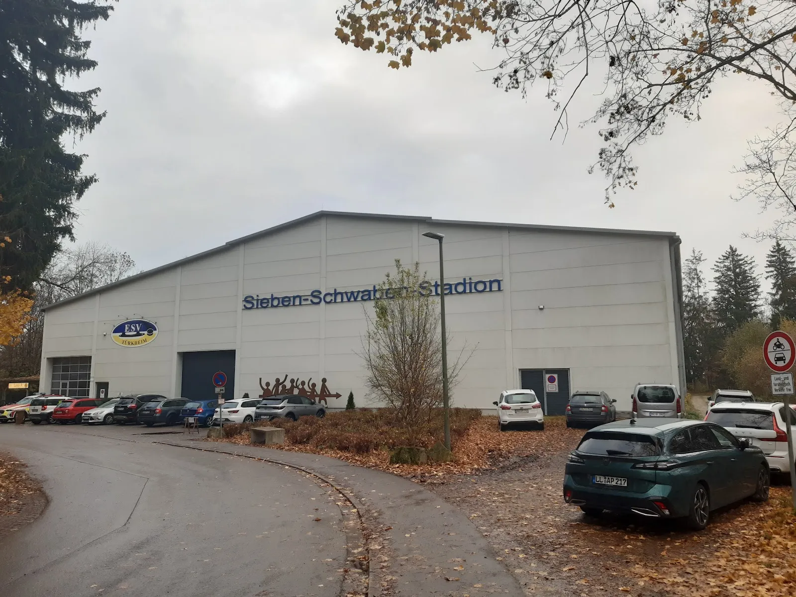 Die große weiße Halle mit dem Schriftzug Sieben-Schwaben-Stadion und dem Vereinswappen, gesehen von der geschwungenen Zufahrtsstraße, mit Autos zwischen Herbstlaub. – Kunsteisstadion Türkheim