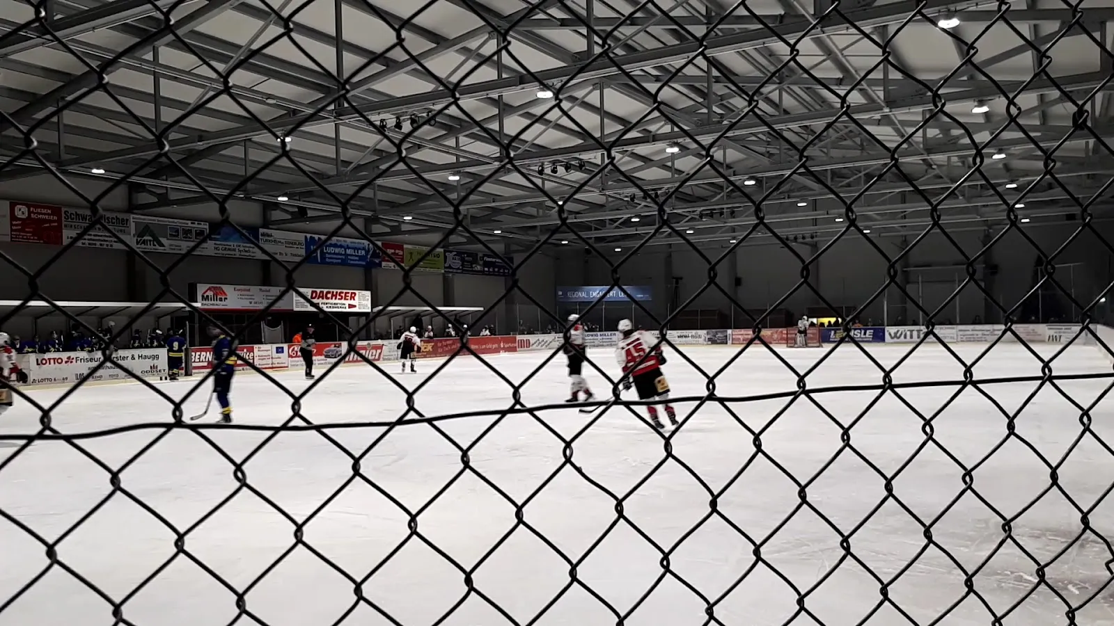 Das Eis und seine Werbebanden, von der Seite der Halle durch einen Maschendrahtzaun gesehen, mit Spielern in der Ferne unter einem beleuchteten Stahldach. – Kunsteisstadion Türkheim