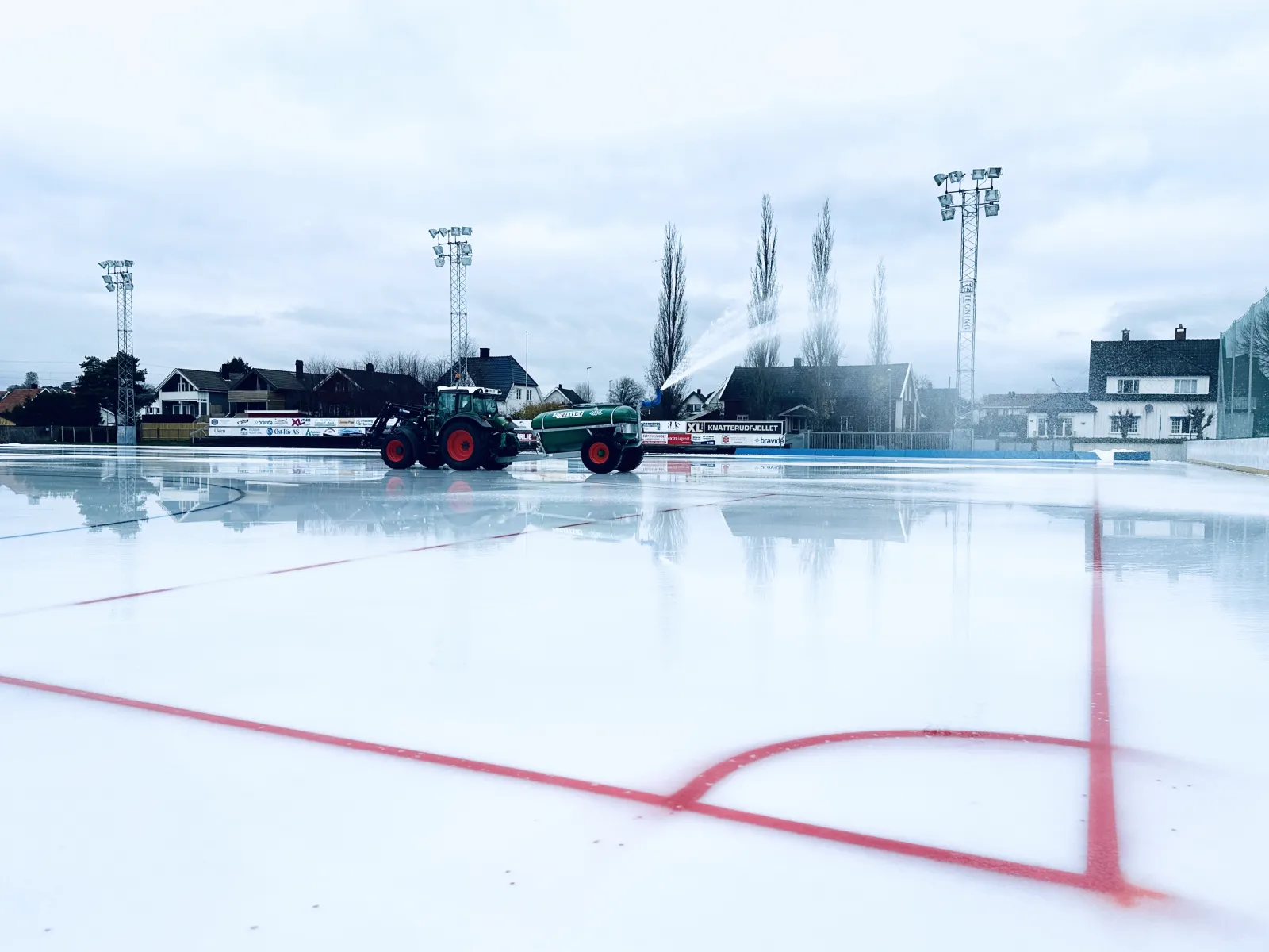 Fresh ice being flooded at the Sarpsborg kunstisbane, with the red lines marked and the surrounding houses and floodlights behind. — Kunstisbanen ved Sarpsborg stadion