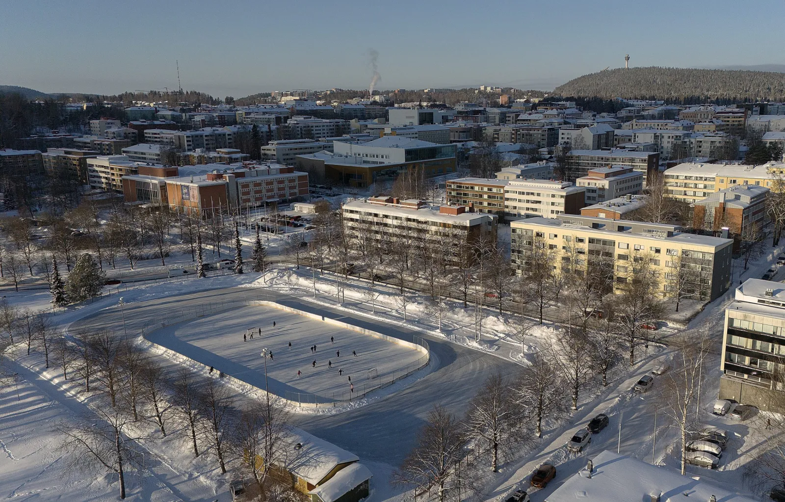 An aerial view of the Kuopionlahti skating oval in Maria Jotuni park, with skaters at a distance and apartment blocks beyond. — Kuopionlahden luistelukenttä