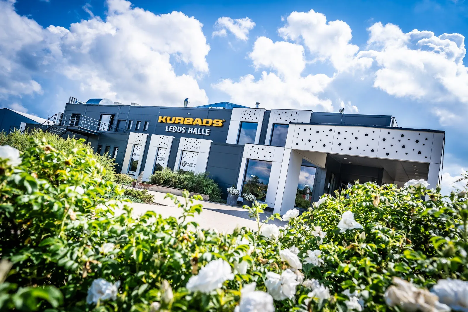 The front of the Kurbads ice hall, a low building with the KURBADS LEDUS HALLE lettering above the entrance and flowering shrubs in the foreground. — Kurbads ledus halle