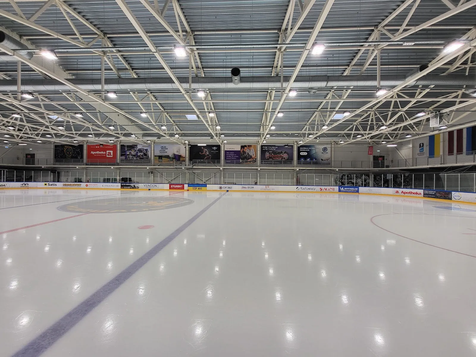 The empty ice sheet of the main hall with the dasher boards, spectator seating and the steel roof trusses. — Kurbads ledus halle