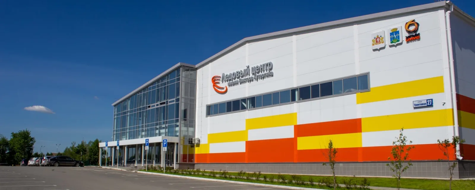 The white, yellow and orange building of the Kutergin Ice Centre in Sysert, with its Ice Centre sign, seen from the car park side under a blue sky — Kutergin Ice Centre Sysert