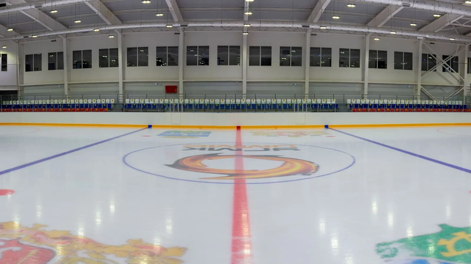The empty ice sheet of the Kutergin Ice Centre seen low from the boards, with the centre circle and red line, the glass above the boards and the roof trusses — Kutergin Ice Centre Sysert