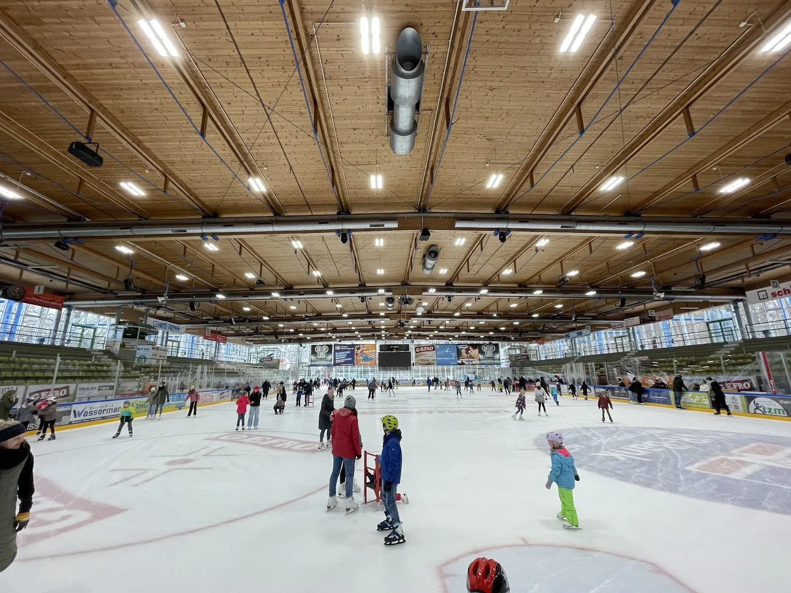 Ein Publikumslauf auf dem Eis der Memminger Eissporthalle (KUTTER-Arena): Schlittschuhläufer über die ganze Fläche, darüber die Holzdecke und hinter der Bande die Ränge. – KUTTER-Arena