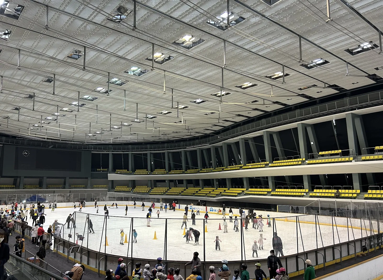 The main rink at Kyoto Aquarena during a public session, with many skaters on the ice and spectators along the boards under a ceiling of skylights. — Kyoto Aquarena
