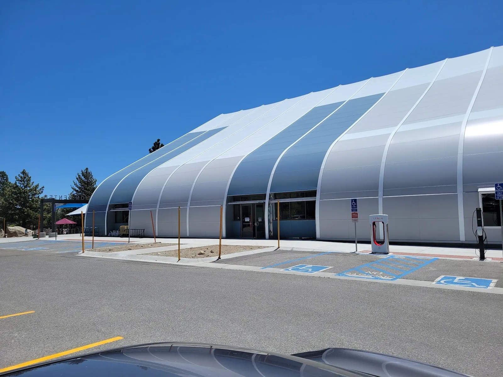 The long white curved tent building of LA Kings Ice at Mammoth Lakes with a glazed front, seen from the car park under a blue sky and pine trees. — LA Kings Ice at Mammoth Lakes