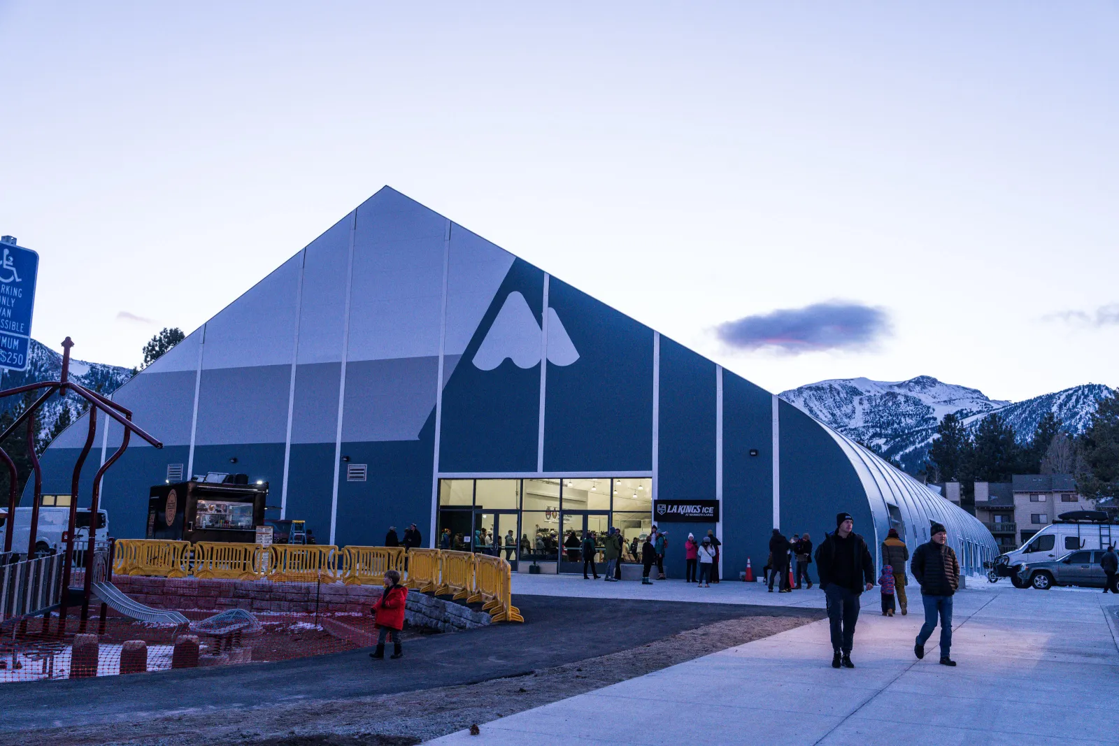 The building's blue gabled facade with its white mountain-peak logo, glass entrance and "LA Kings Ice" sign, photographed at dusk with the Sherwin Range behind it; a few visitors are gathered near the doors. — LA Kings Ice at Mammoth Lakes