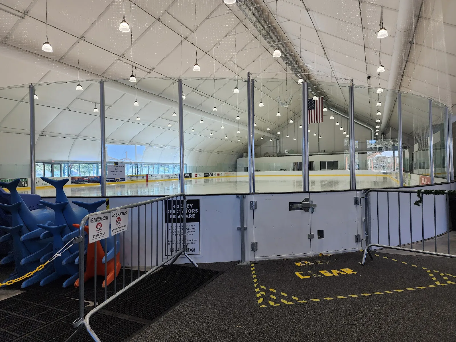 The empty ice sheet seen across the boards and glass inside the arched white fabric building, with a flag on the far wall, seat benches behind and a cleared yellow-marked entrance gate in the foreground. — LA Kings Ice at Mammoth Lakes