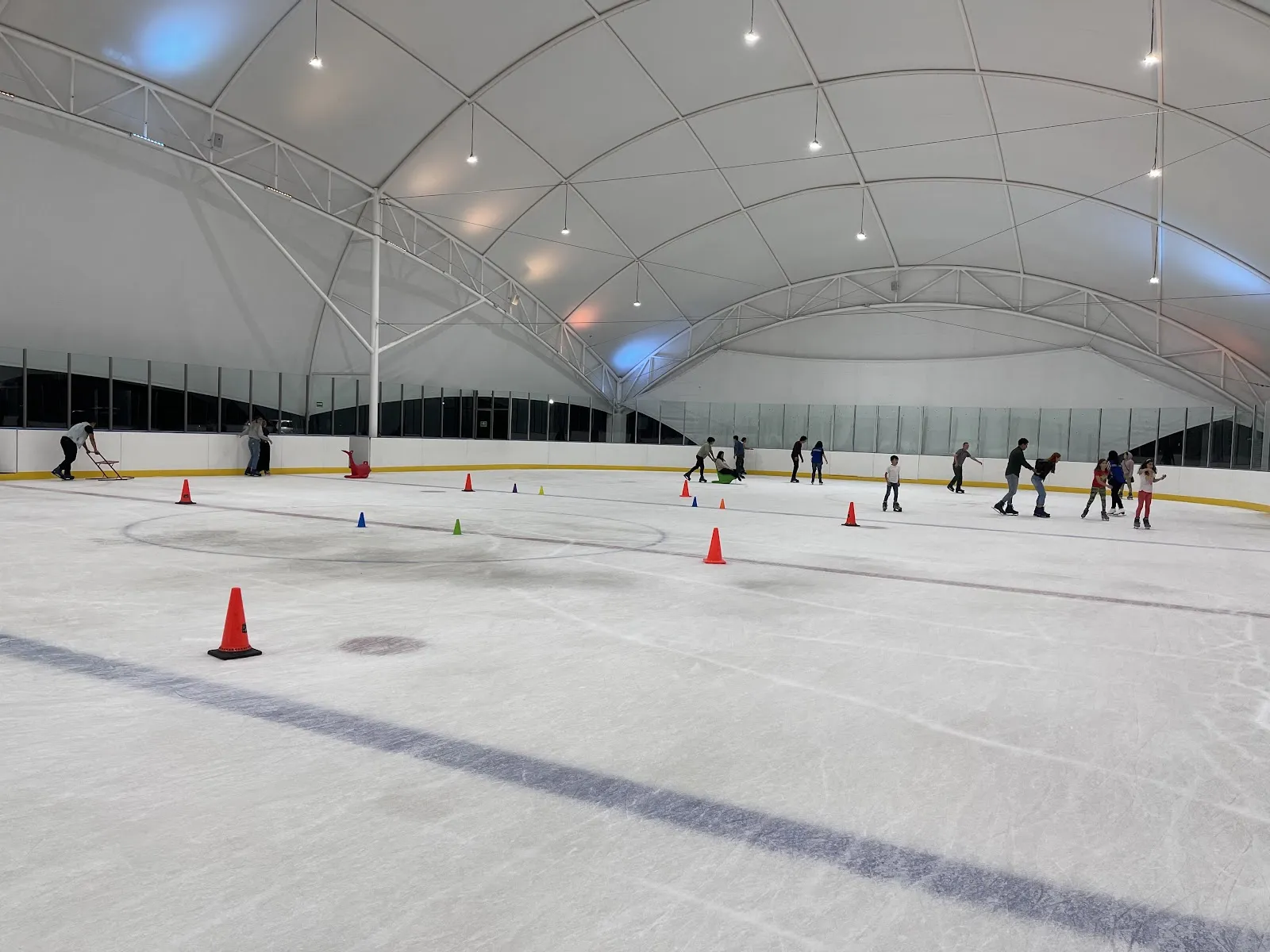 Pista de hielo bajo un domo de lona blanca sobre arcos de acero, con conos de entrenamiento en el hielo y patinadores junto a las vallas del fondo. — La Pista Coyoacán