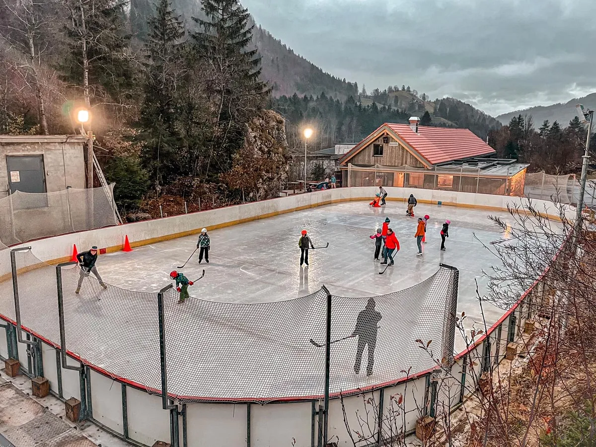 La Scie outdoor ice rink at Charmey: a boarded rink lined with netting, skaters spread across the ice, a wooden chalet-style building and wooded mountain slopes behind. — La Scie Patinoire de Charmey