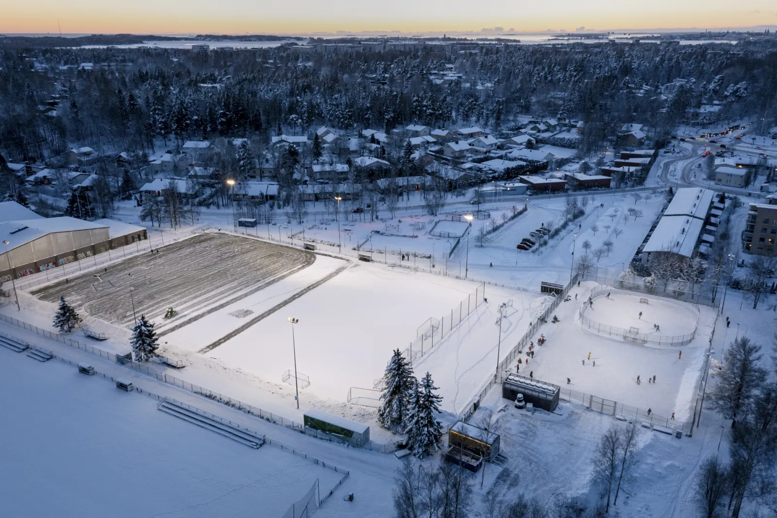 People skating on the ice rink in Laajasalo Sports Park, which has a hockey rink on one side. — Laajasalon liikuntapuisto – tekojääkenttä