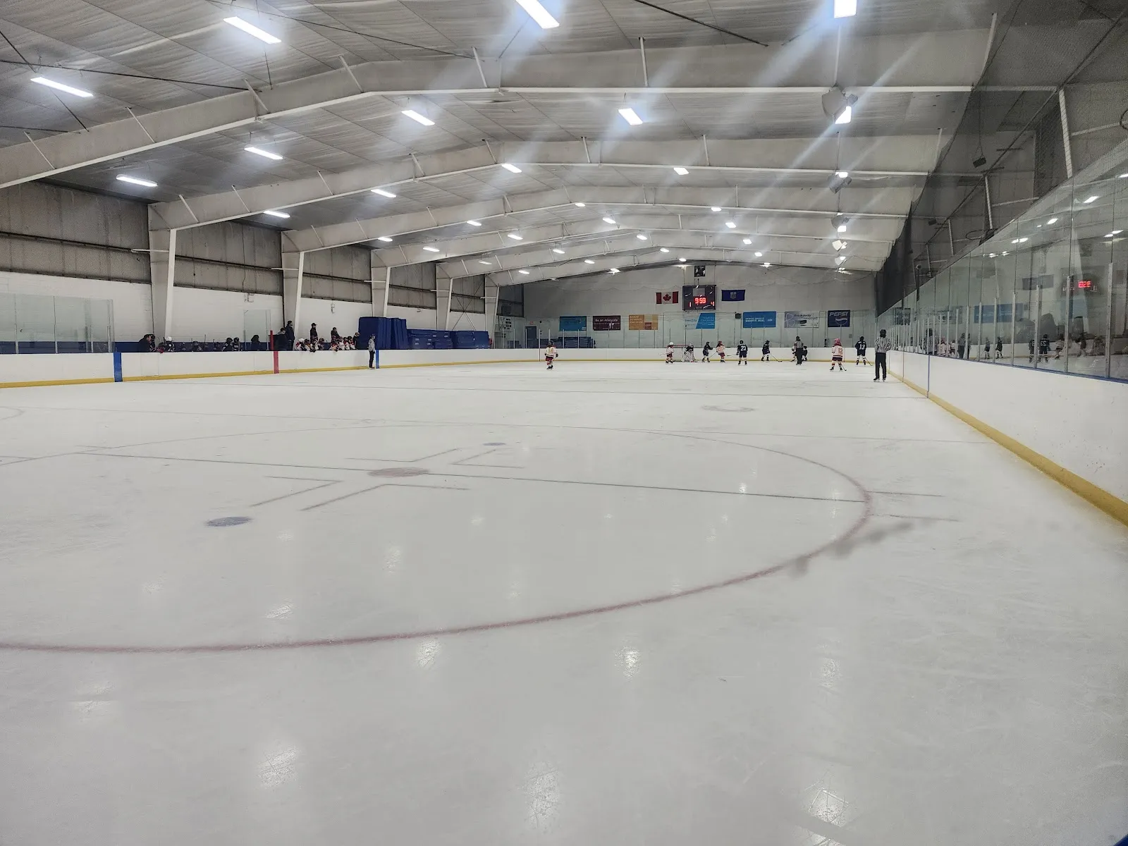 The rink from centre ice under a bright white roof, with players small at the far end. — Labour Club Ice Centre