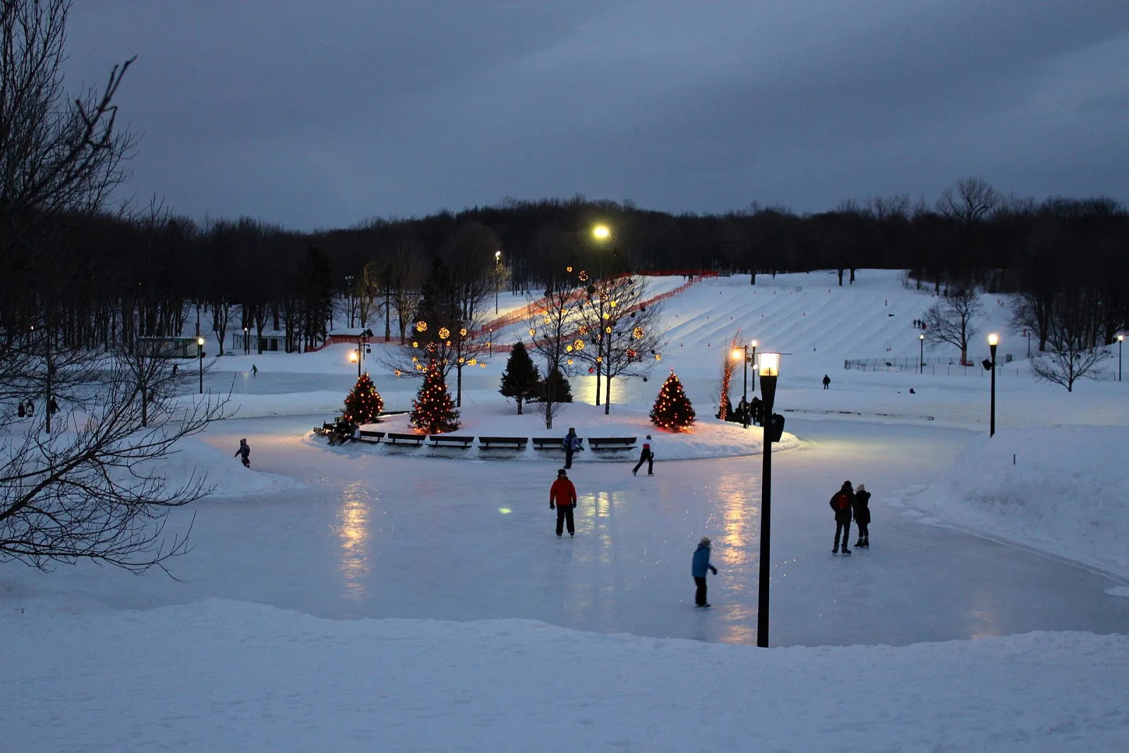 The Beaver Lake skating rink on Mount Royal at dusk, lit by lamps and Christmas trees, with a few skaters on the ice — Lac aux Castors Rink (Parc du Mont-Royal)