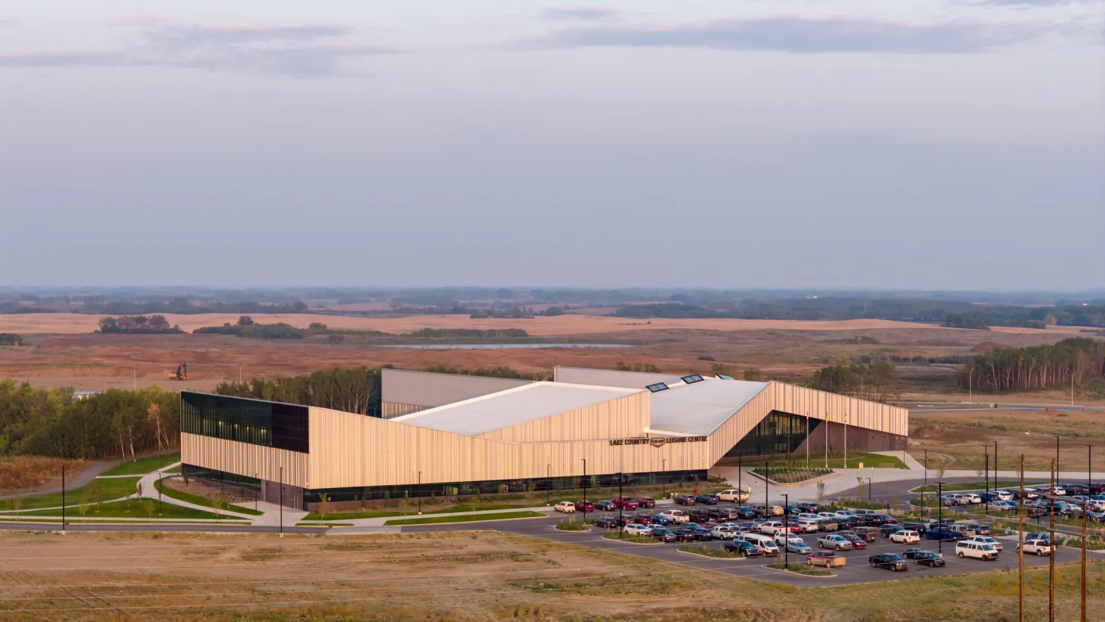 El Lake Country Co-op Leisure Centre desde arriba al atardecer: techos angulares de revestimiento claro sobre muros de vidrio, un estacionamiento lleno y la pradera al fondo. — Lake Country Co-op Leisure Centre