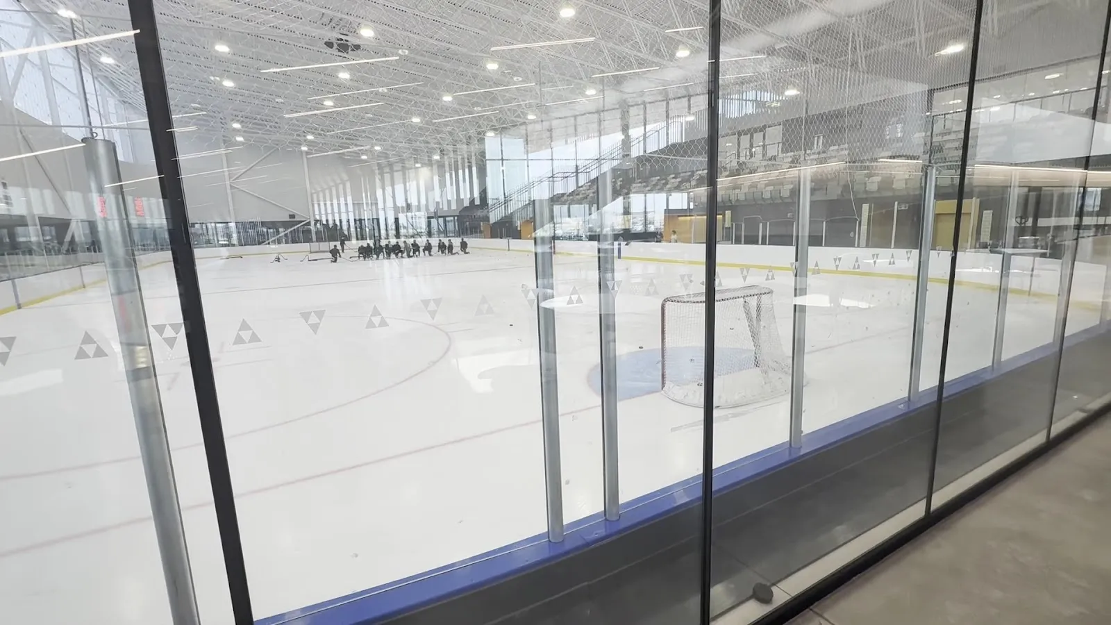 Un panel de vidrio transparente que da a una pista luminosa y vacía, con un techo con estructura de acero y una pared de ventanas más allá de las vallas. — Lake Country Co-op Leisure Centre
