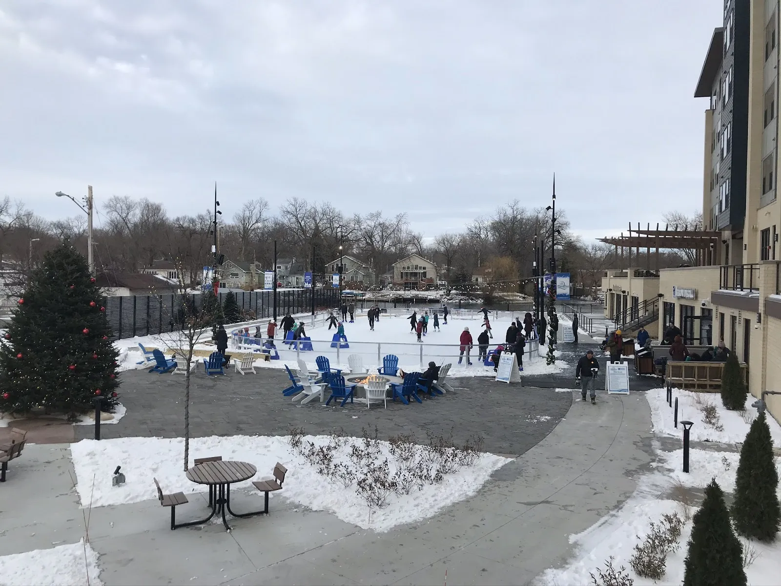 The plaza and fire-pit seating beside the Grand Crossing Park rink in Monona, with apartment buildings behind — Lake Ridge Bank River Rink