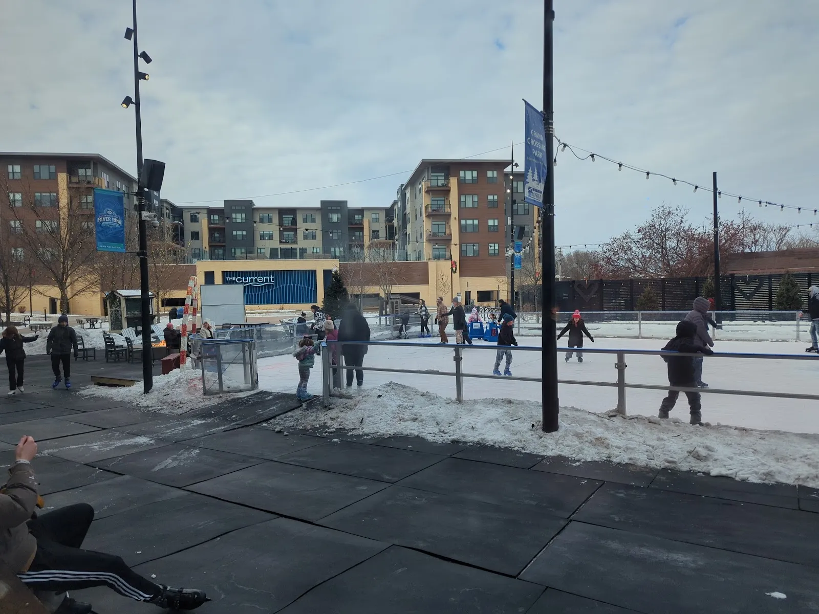 Skaters seen from a distance on the plaza rink in daylight, with the rink boards and light poles — Lake Ridge Bank River Rink
